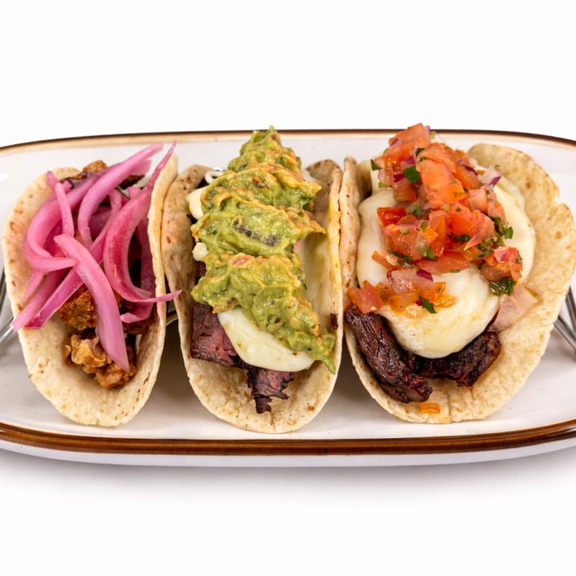 Three A Fuego Lento tacos with various toppings, including pickled onions, guacamole, and salsa.