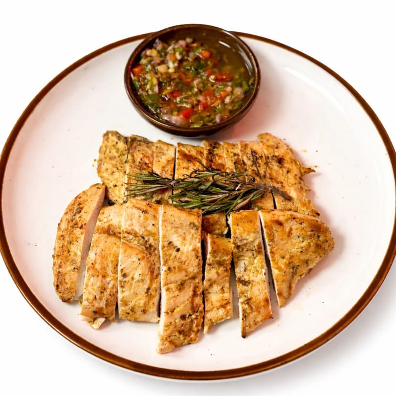 Sliced grilled chicken breast with rosemary sprigs and a side of chimichurri sauce on a white plate.