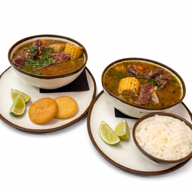 Two bowls of A Fuego Lento's beef soup with corn, cilantro, lime, and a side of rice or fried dough.