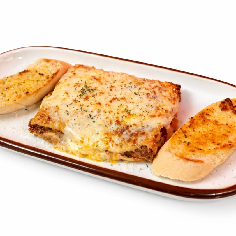 A Fuego Lento's cheesy lasagna with two slices of garlic bread on a white rectangular plate.
