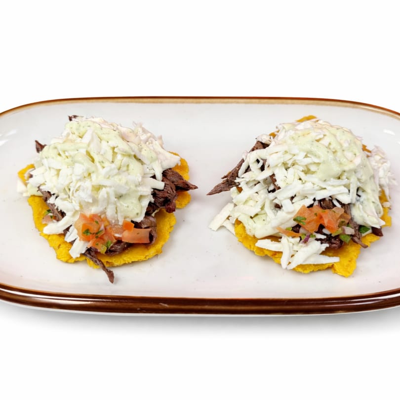 Two A Fuego Lento tostones topped with shredded beef, white cheese, green sauce, and pico de gallo.