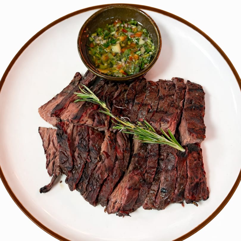 Sliced grilled steak with a sprig of rosemary and a side of chimichurri sauce on a white plate.